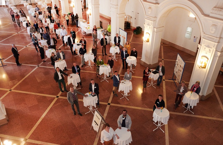 Gruppenfoto der etwa 60 Konferenz-Teilnehmenden in einem festlichen Saal.  Jeweils zwei Personen stehen an einem Stehtisch.